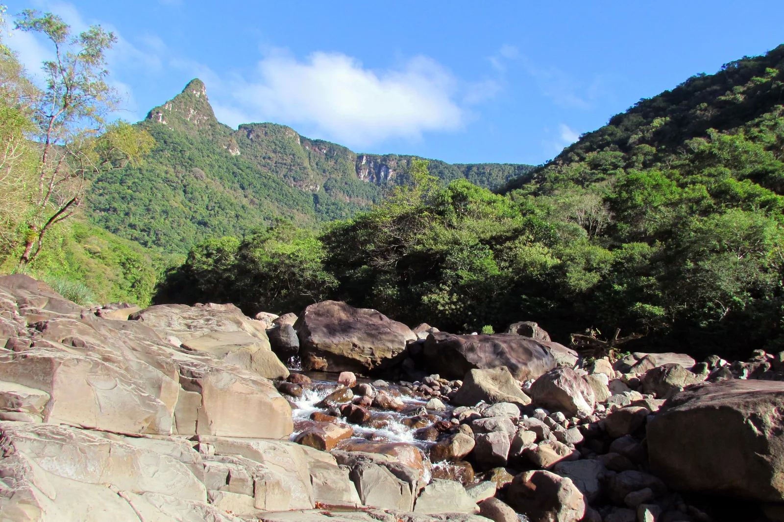 Pale boulders in a rocky riverbed at the start of the Rio do Boi trail, a pointed forested peak standing above the trees behind