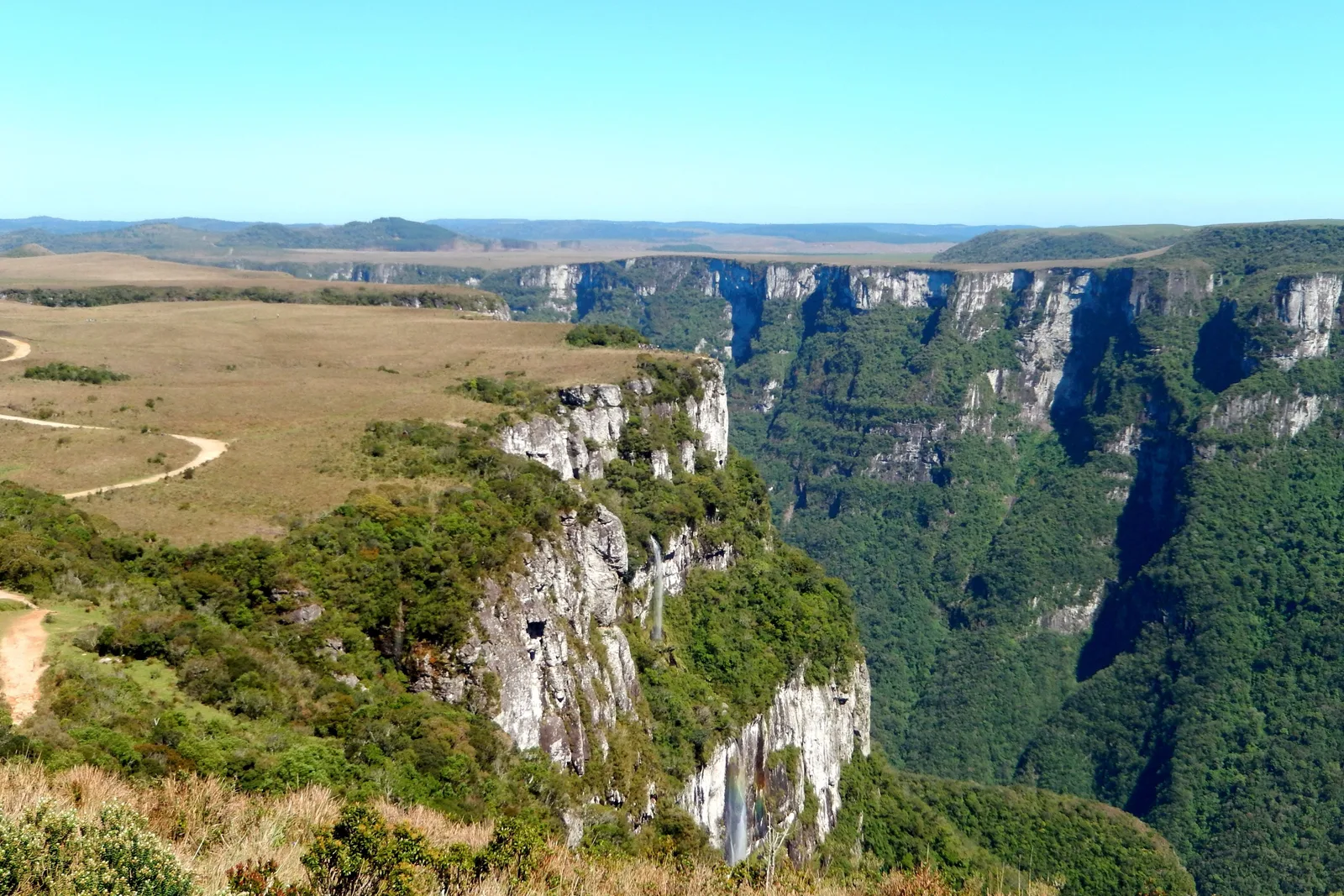 The rim of the Fortaleza canyon, flat yellow grassland and a dirt track running to the edge of forested cliffs that fall away into a long green valley