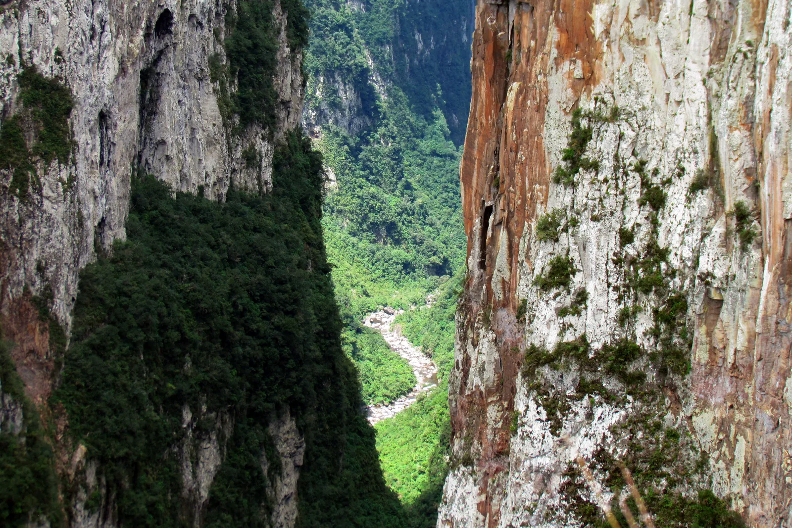 Looking down a narrow gap between two red and gray rock walls in the Itaimbezinho to a river winding along the canyon floor far below