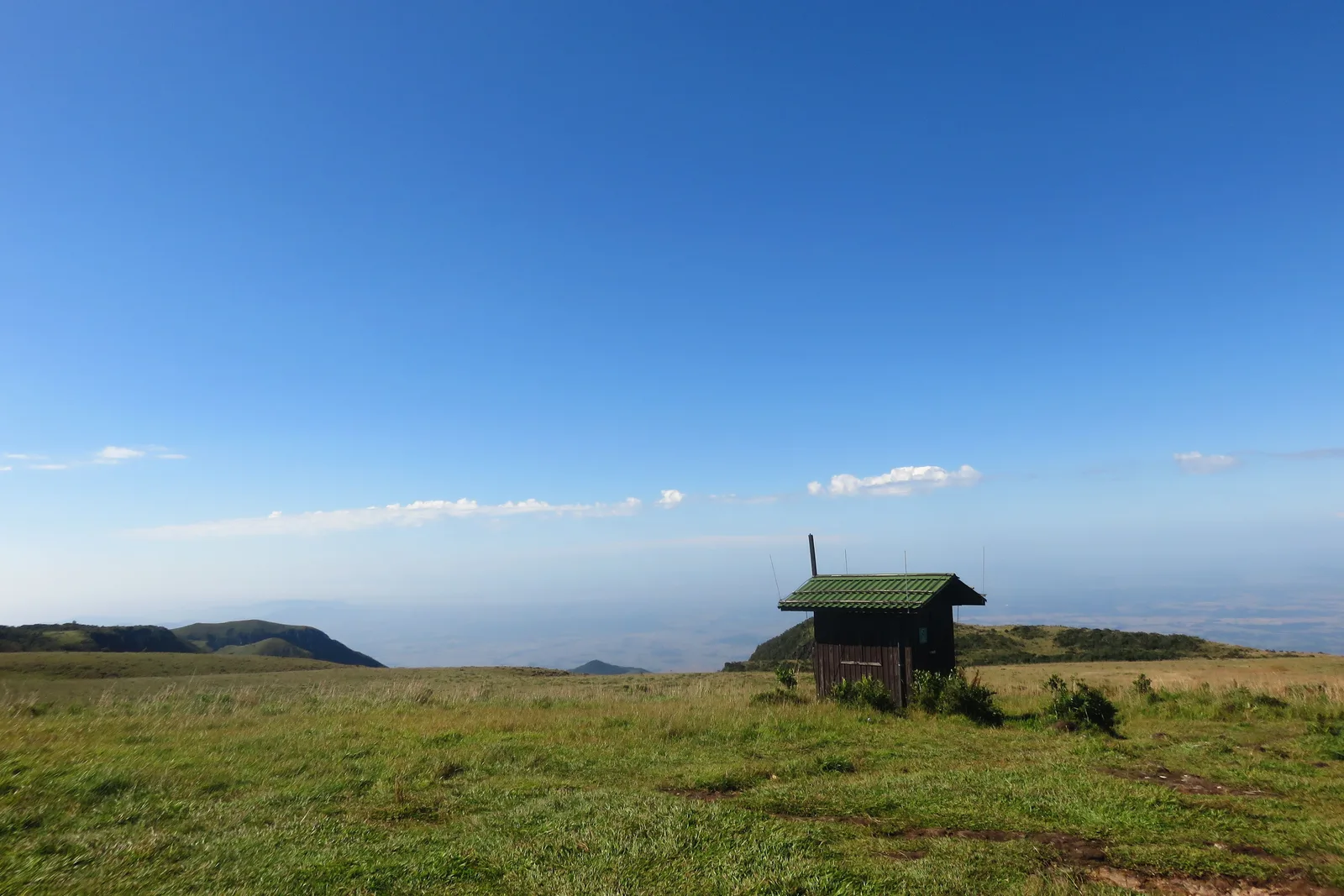 A small wooden hut alone on open grassland at the top of the escarpment, the coastal plain hazy far below under a deep blue sky
