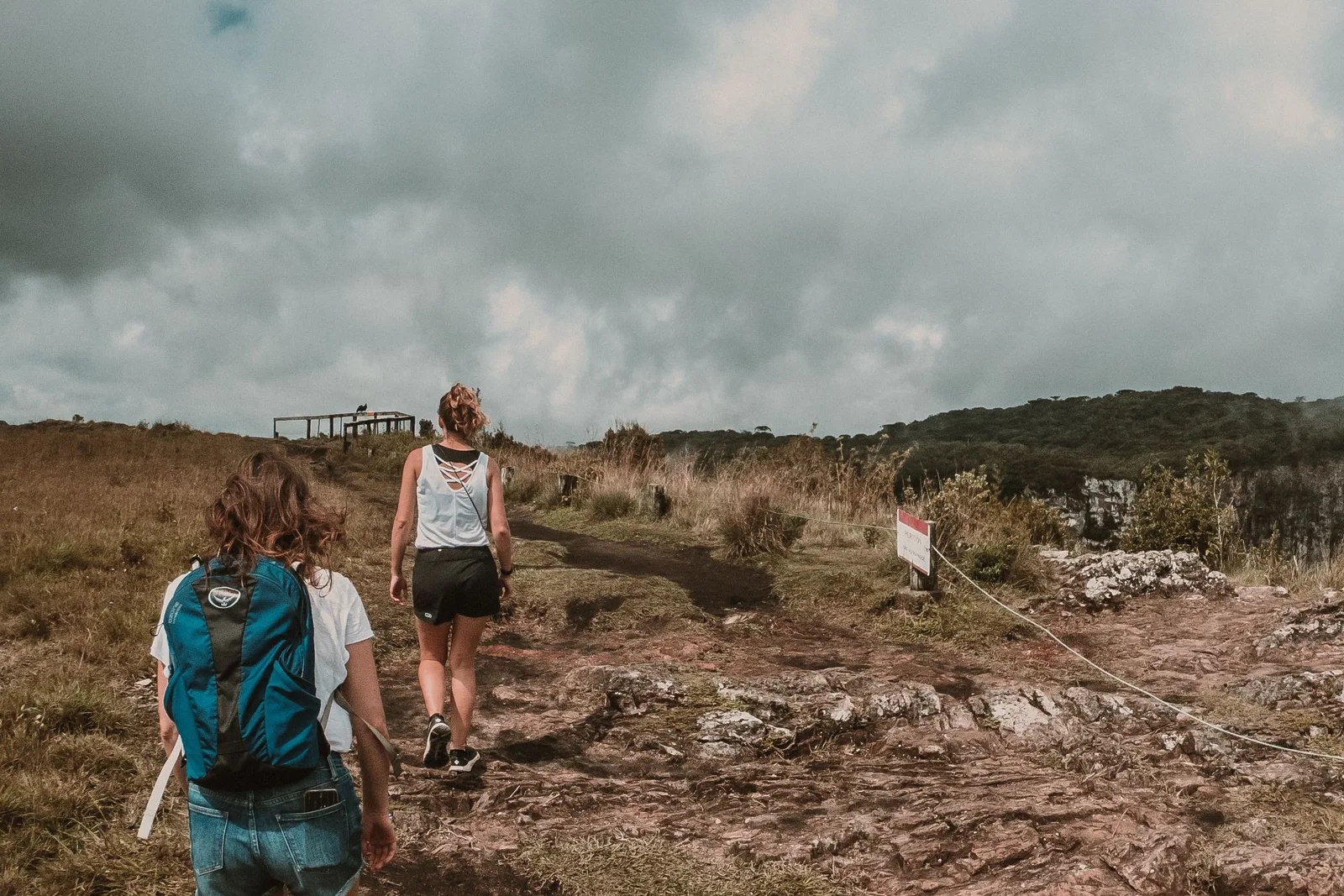 Two hikers, one with a teal daypack, walking across rough dry ground toward a canyon rim under a heavy gray sky, a wooden viewing platform ahead and a rope line beside the path