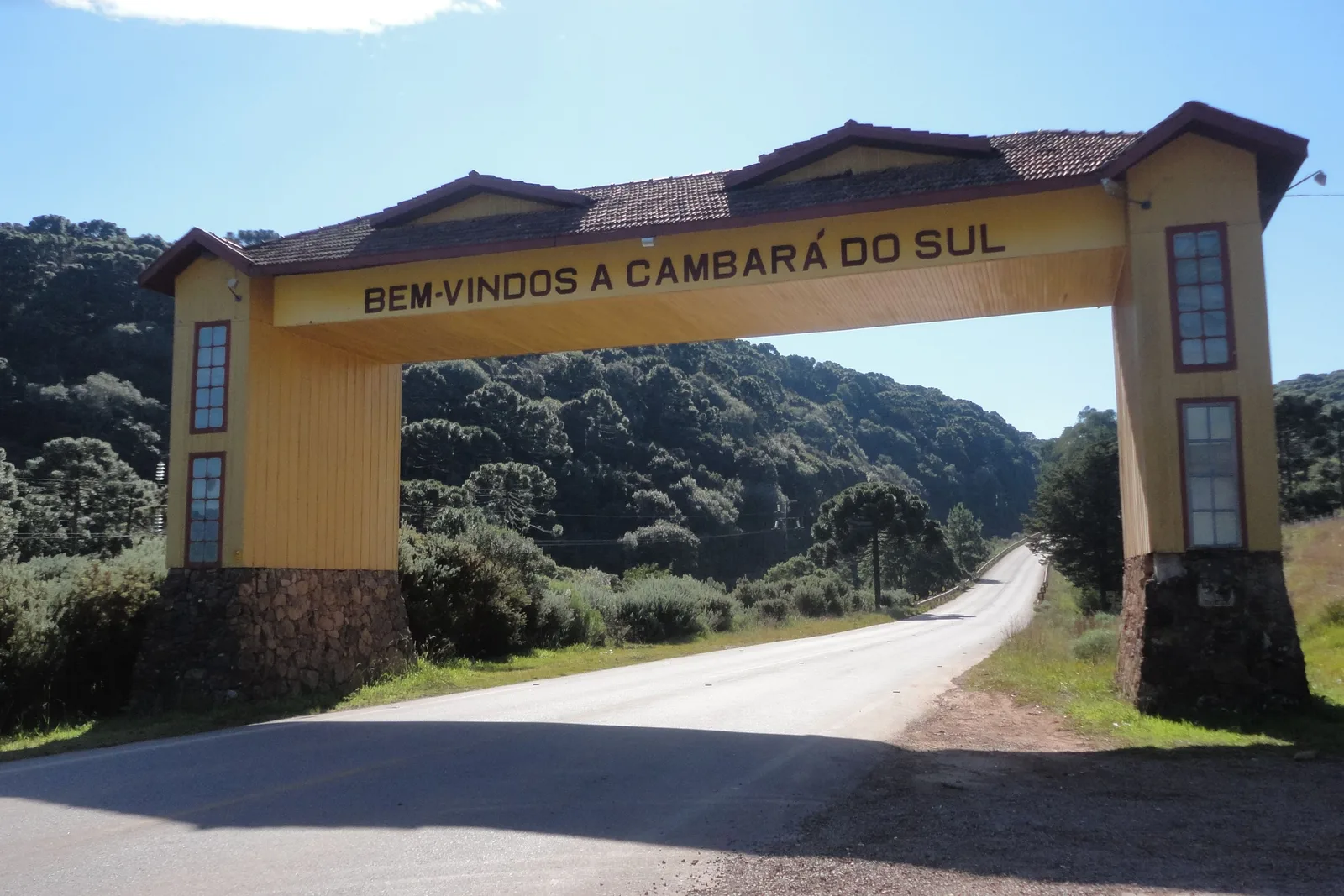 A yellow wooden gateway arch reading “Bem-vindos a Cambará do Sul” over an empty road, araucaria-covered hills behind