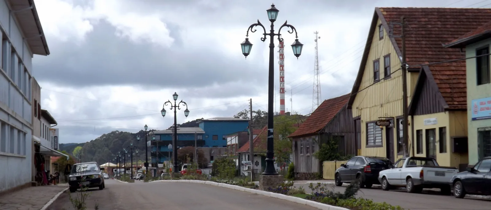 The main street of Cambará do Sul under a gray sky, ornate street lamps down the median and old wooden houses with steep roofs on the right