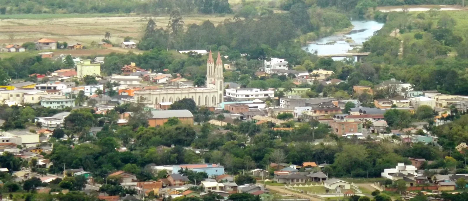 Praia Grande seen from the escarpment above, rooftops and a church with two pointed towers among trees on the plain, a river and green fields beyond