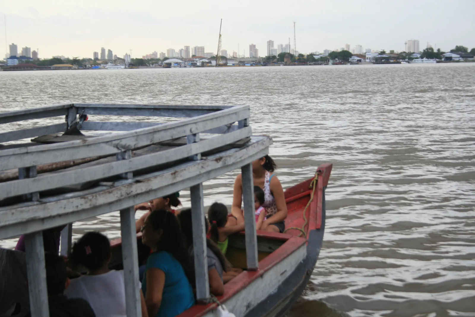 People sitting in a red canoe tied alongside a weathered wooden jetty on a Combu channel, the low skyline of Belém across the open water behind