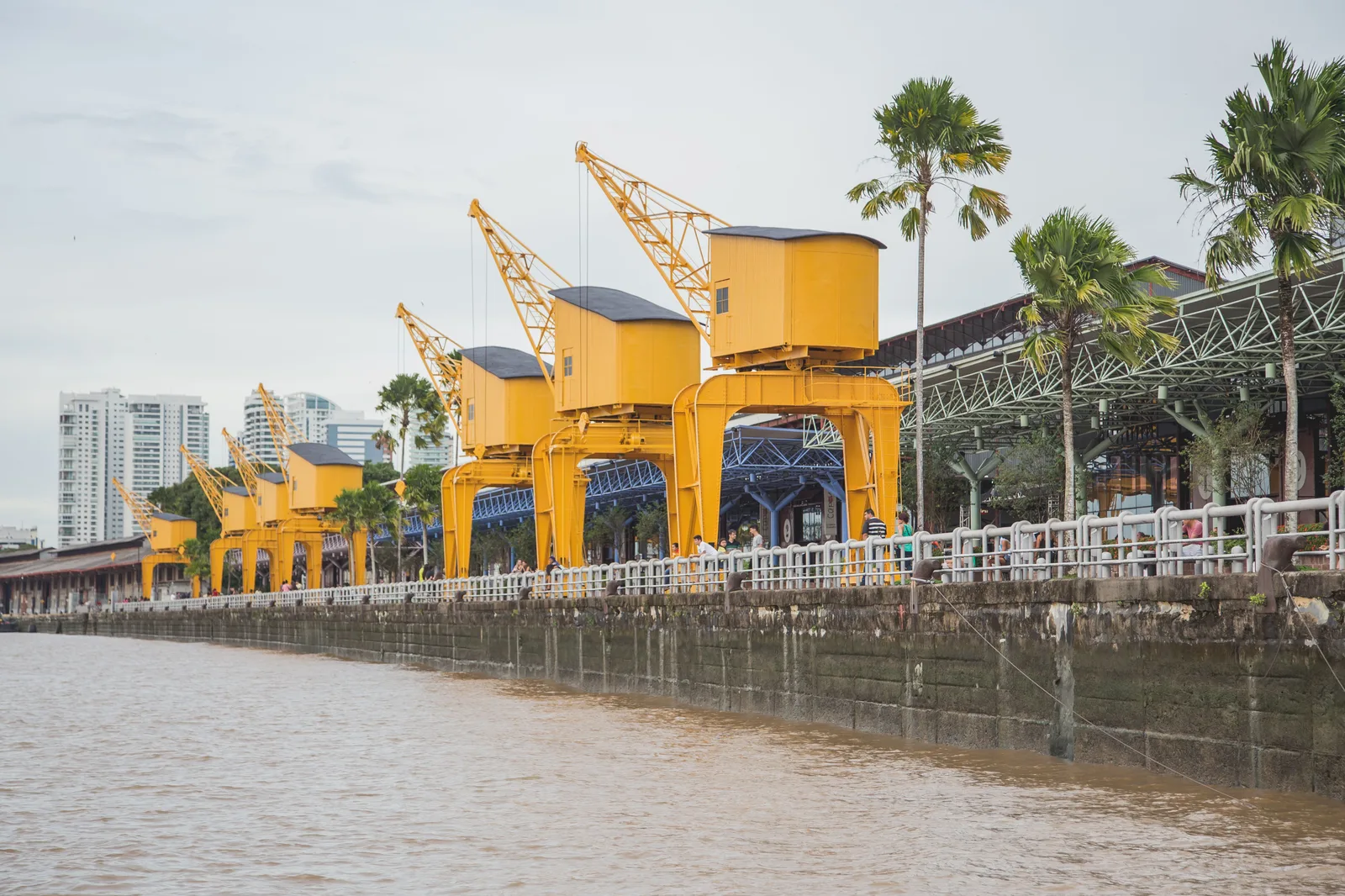 The Estação das Docas from the water: a row of five yellow gantry cranes standing along a quay in front of long steel-framed warehouses, city towers rising behind