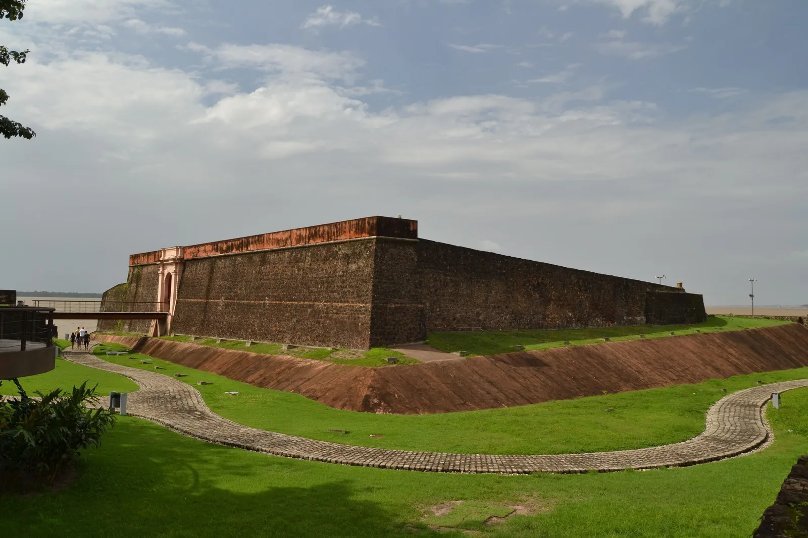 The Forte do Presépio from outside: a low fort of dark stone and brick with sloped grass earthworks running up to its walls, trees behind and a bright sky above