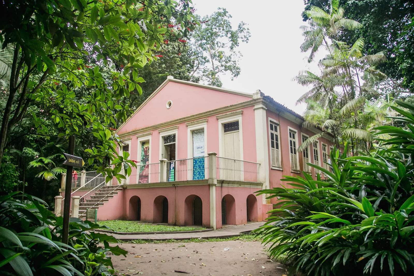 A pink colonial building with a shaded ground-floor arcade standing among palms and dense tropical planting inside the Museu Goeldi park