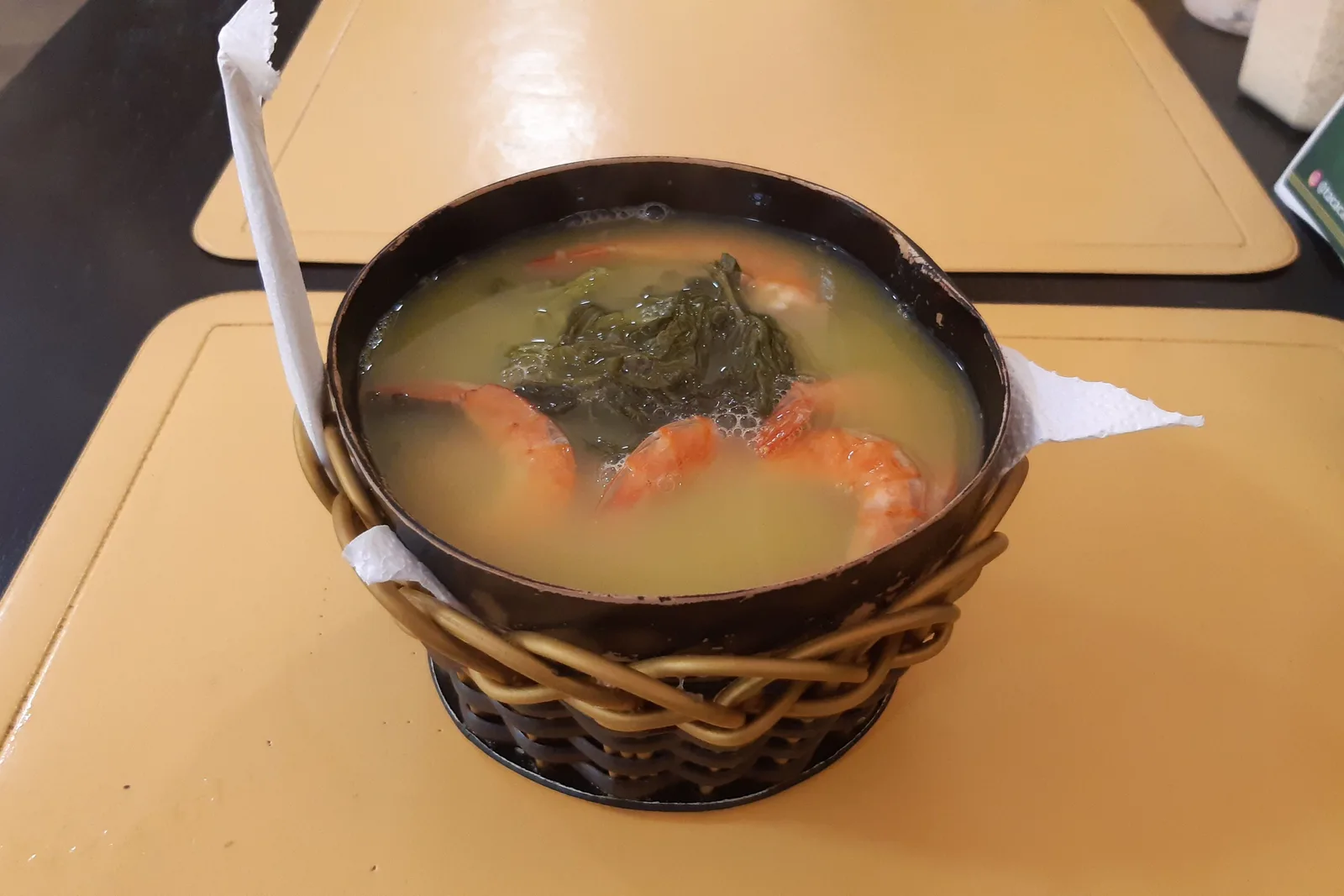 A black bowl of tacacá set in a woven ring on a café table, a paper napkin beside it: pale yellow broth with cooked shrimp round the edge and a clump of dark green jambu in the middle