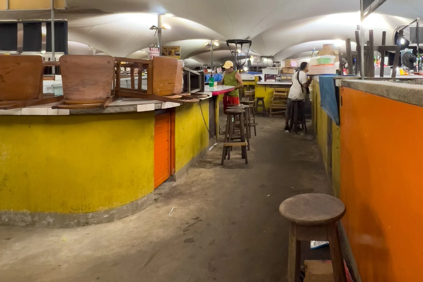 A long curved food counter inside the Ver-o-Peso market painted bright yellow and orange, wooden stools set along it and the market’s vaulted roof overhead