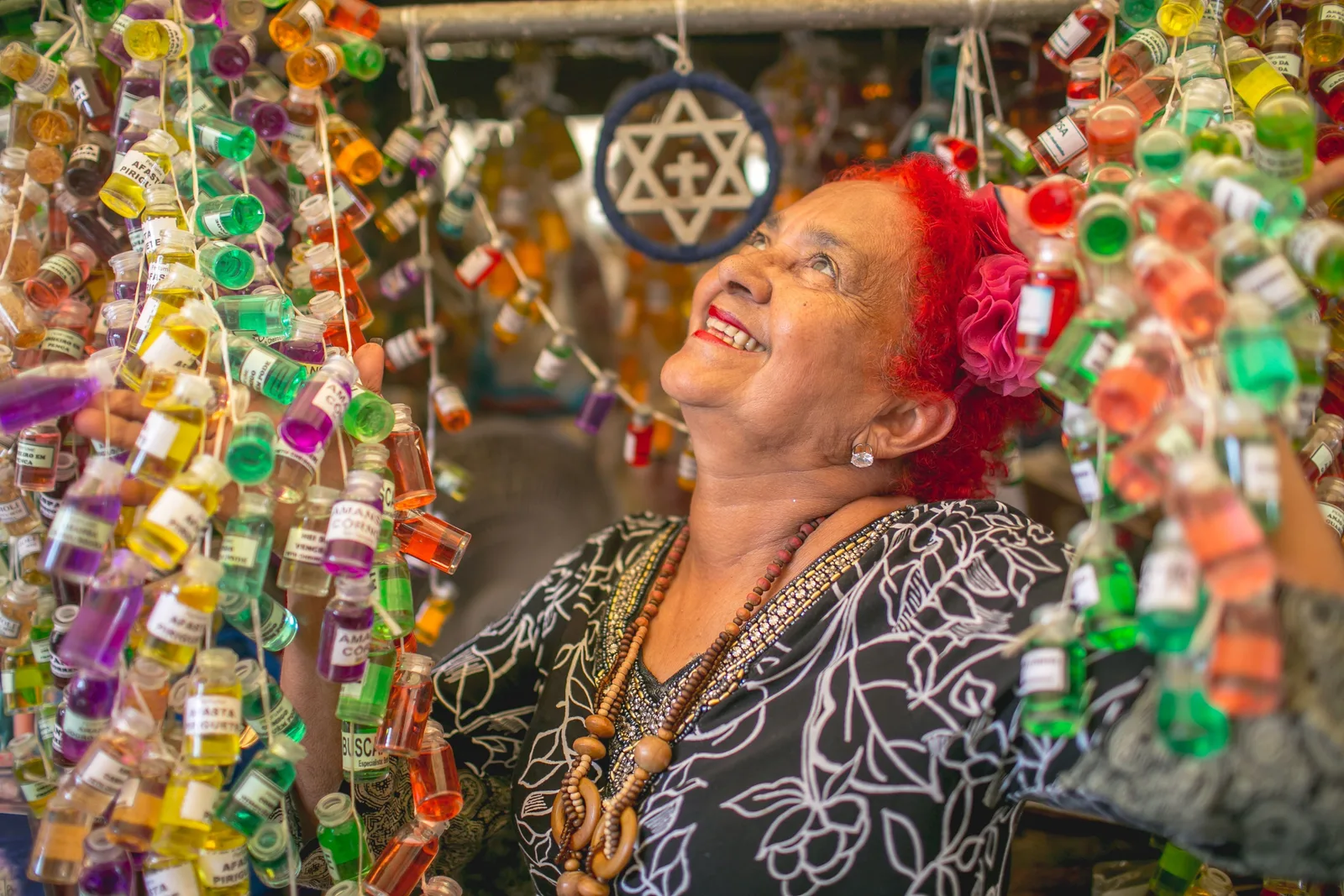 A woman in a patterned blouse with a red flower in her hair laughing beneath dozens of small bottles of colored liquid hung in rows across her market stall