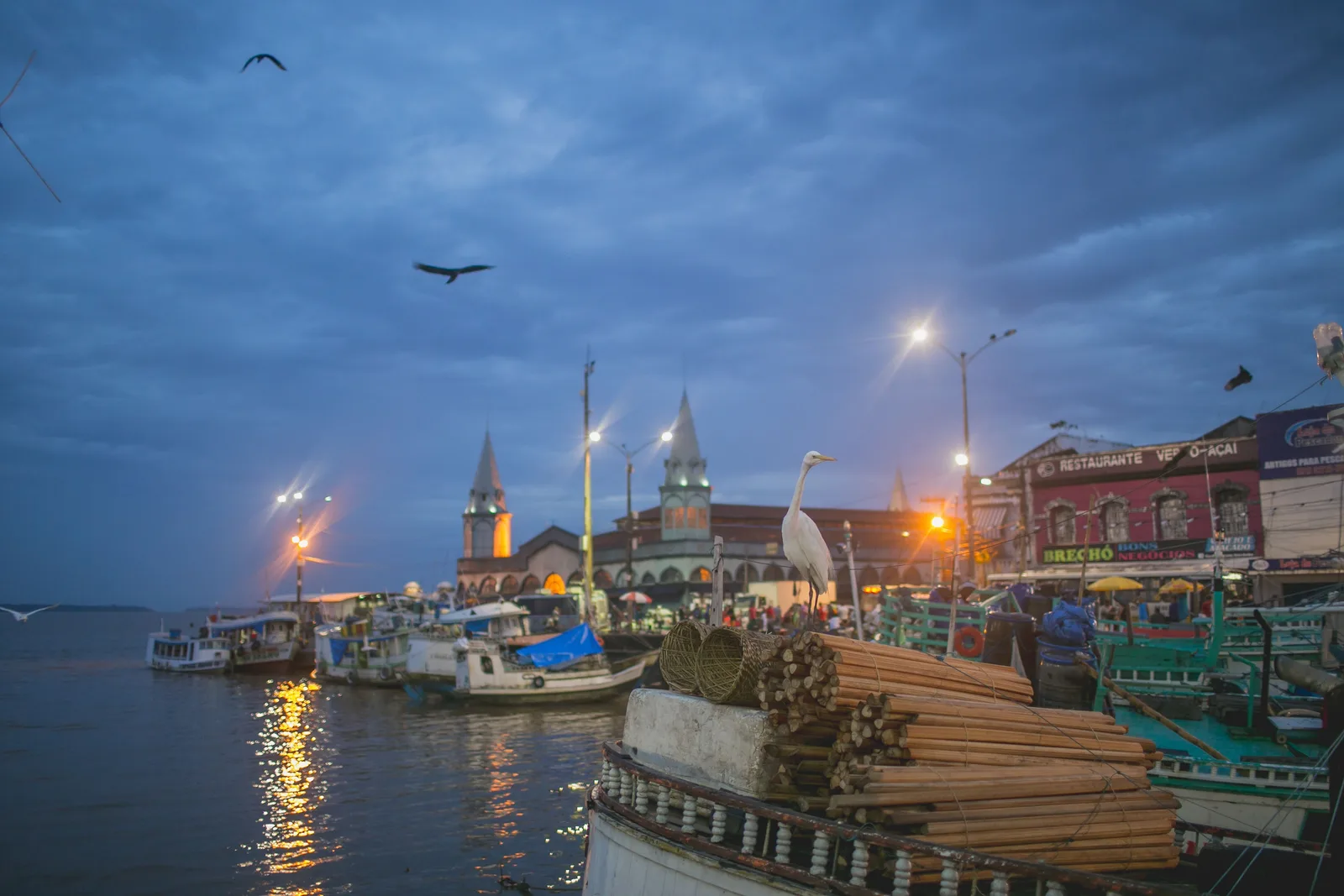 The Ver-o-Peso waterfront at dusk: the market pavilion and its lit corner turret behind a row of moored wooden boats, birds over a deep blue sky