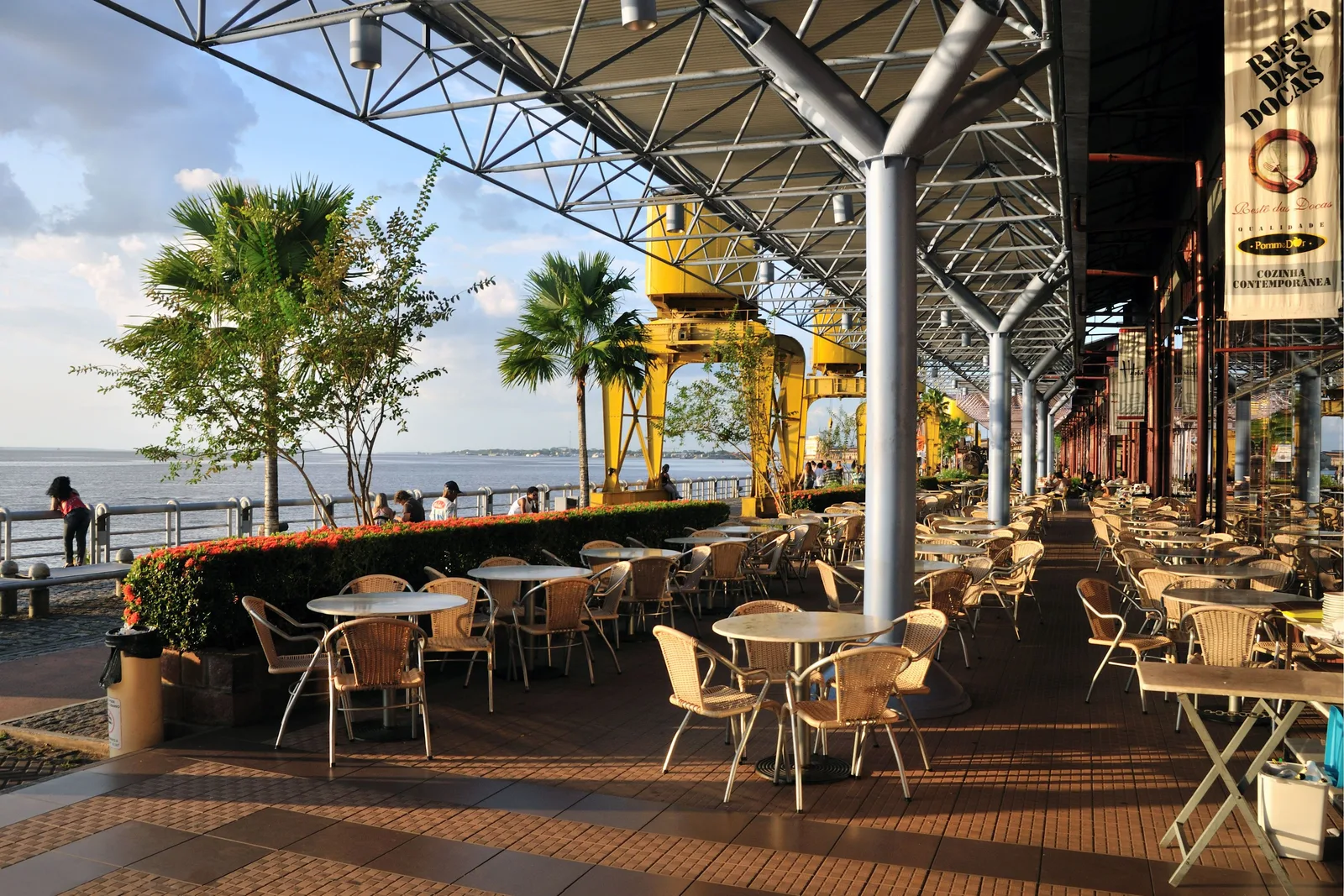 An open riverside terrace under the steel roof of the Estação das Docas, round tables and wicker chairs set out along the railing with the wide pale bay beyond
