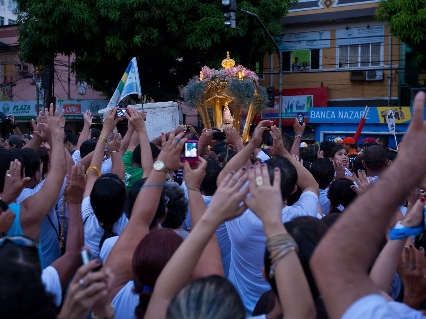 A packed street during the Círio: hundreds of people with arms raised and phones held up, the flower-covered carriage just visible above the crowd in the distance