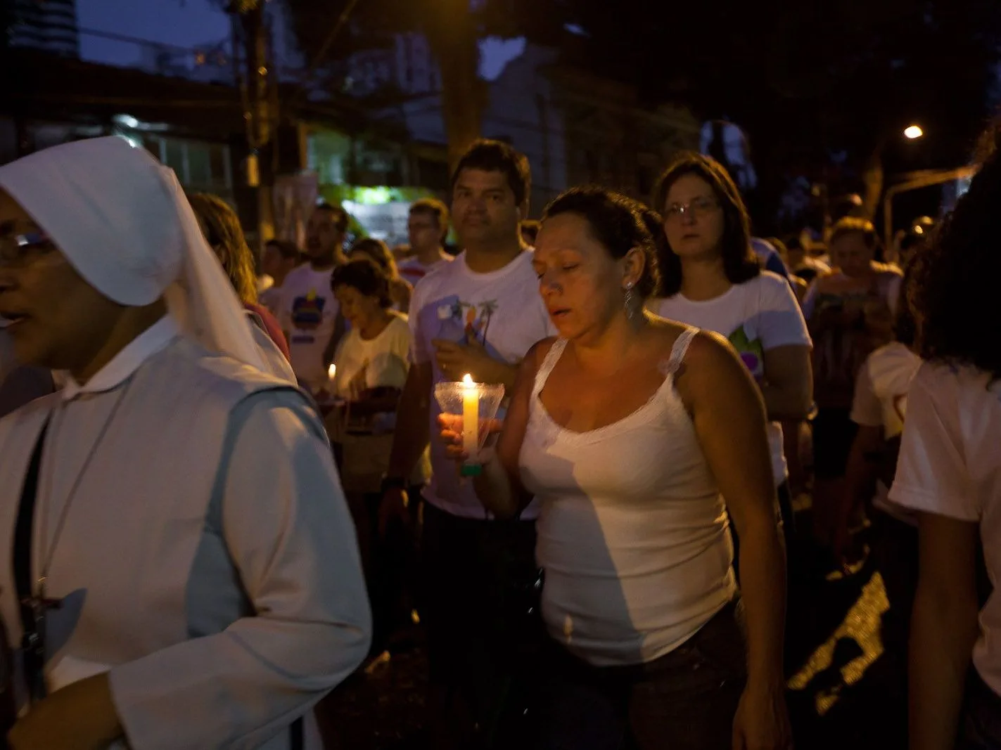 A night procession in Belém: a nun in a white habit and people in white shirts walking closely together, several holding lit candles, in a lamplit street