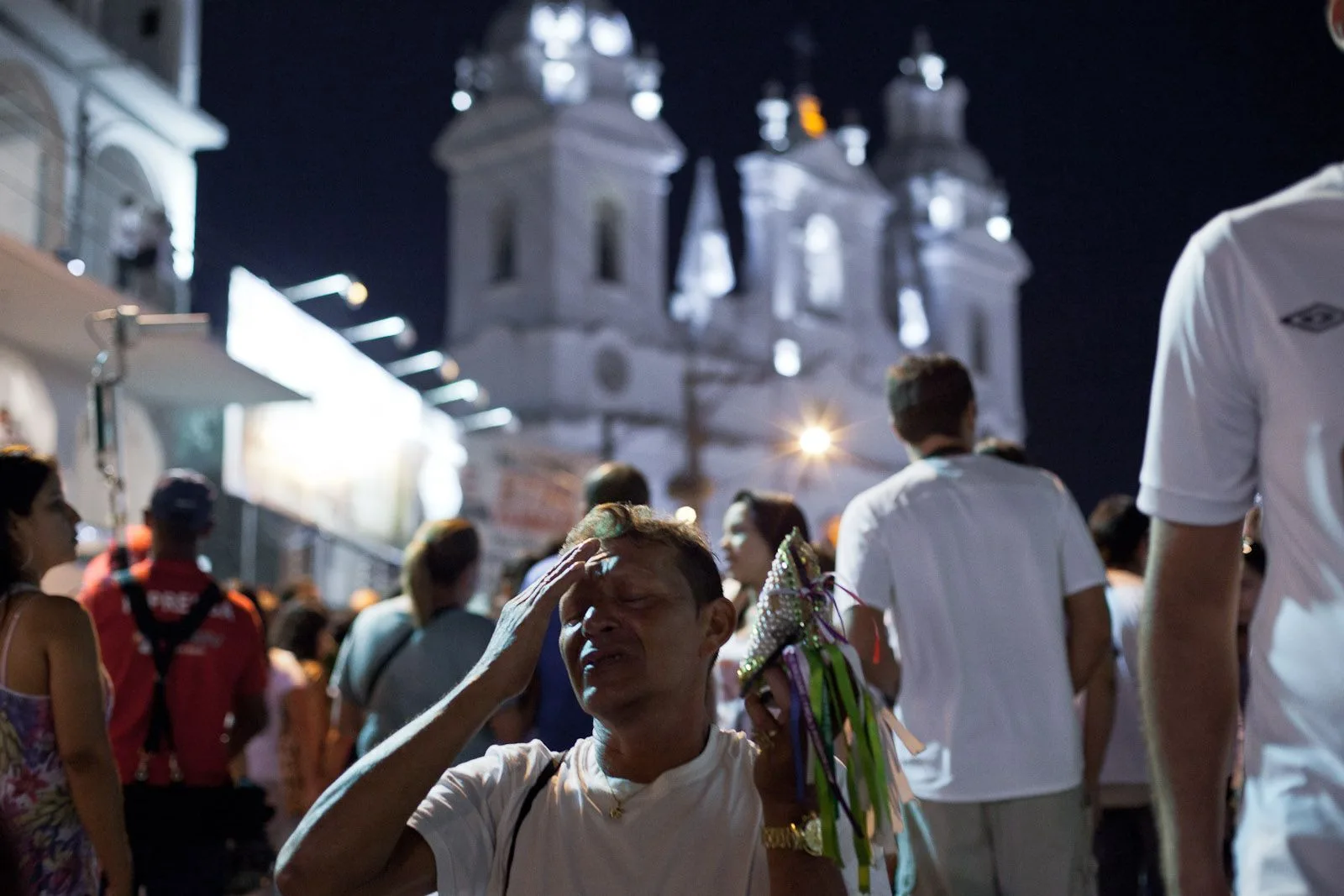 A floodlit white church with lit towers rising behind a crowd of people filling a street at night during the Círio