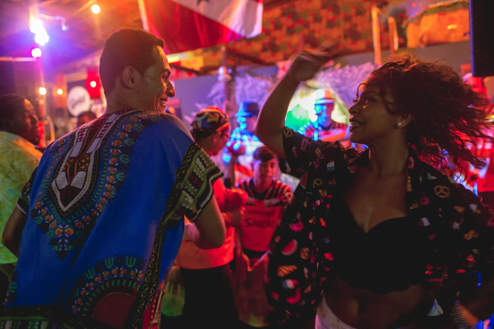 Two dancers in bright printed clothes moving under colored lights at a night party in Belém, a crowd behind them
