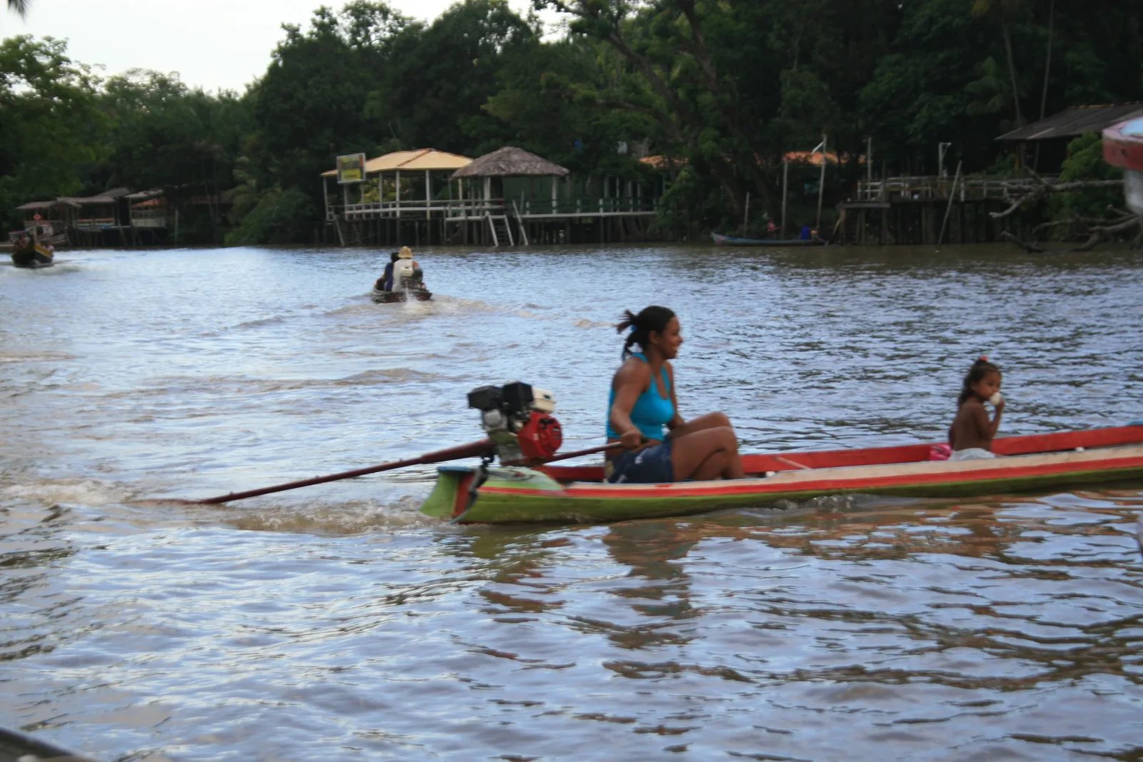 A family sitting in a narrow red canoe on the brown water off Ilha do Combu, wooden houses on stilts along the bank behind them