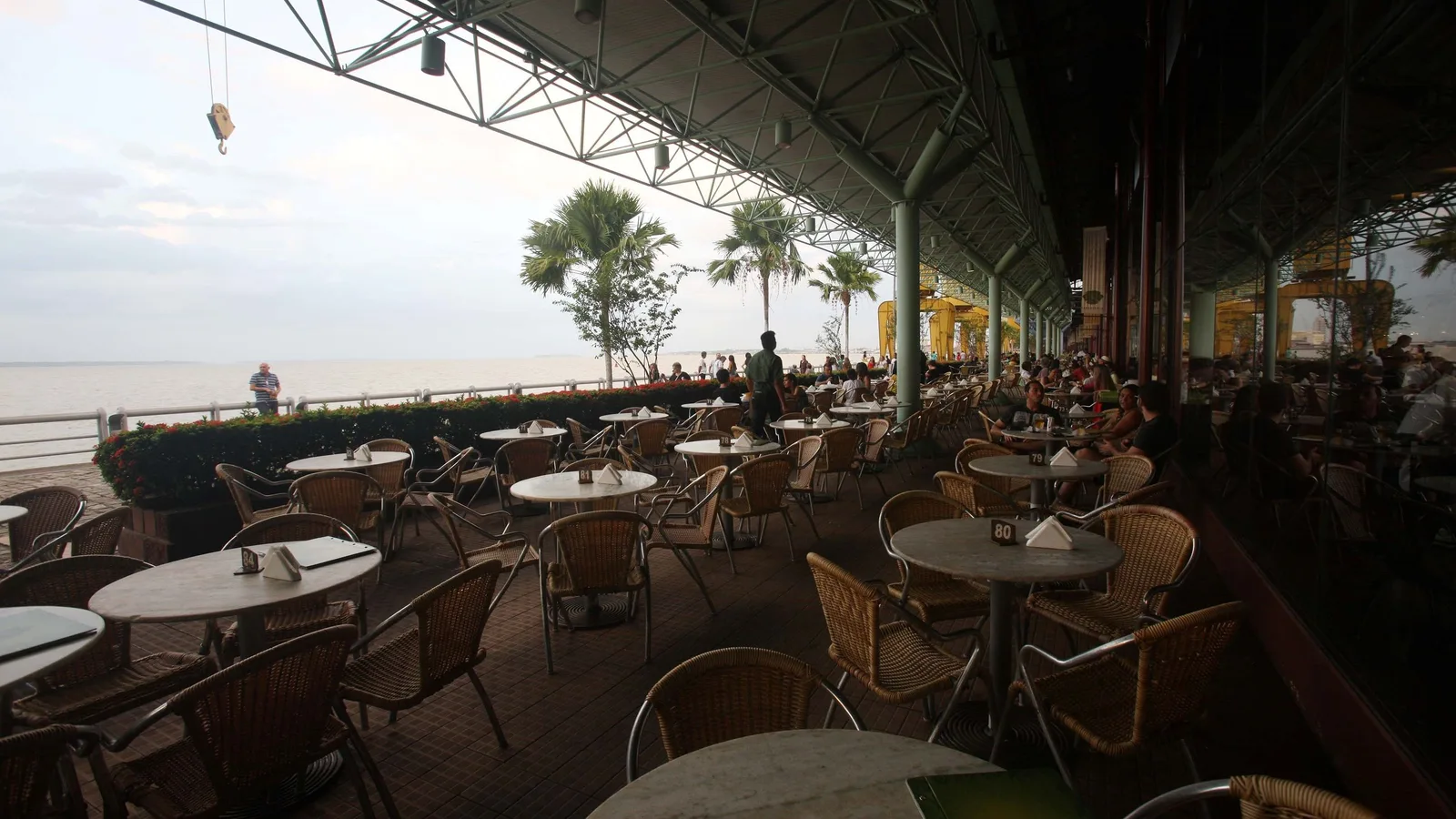 The riverside terrace of the Estação das Docas at dusk, tables and chairs set out along the railing under the warehouse roof with the bay going dark beyond