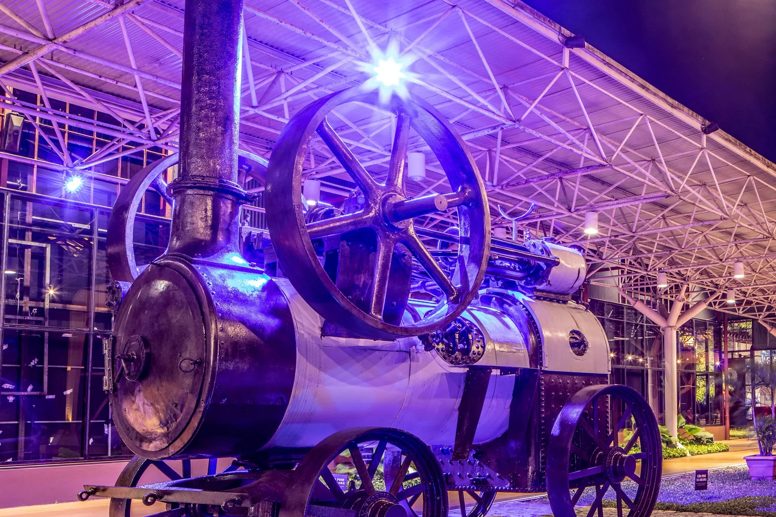 An old riveted steam engine on iron wheels displayed under the warehouse roof at the Estação das Docas, floodlit deep blue and violet