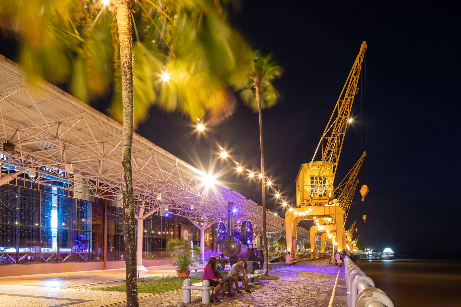 The Estação das Docas at night: a tall yellow dockside crane lit from below beside a long steel-framed warehouse, palms and a lit promenade running along the water in front of it