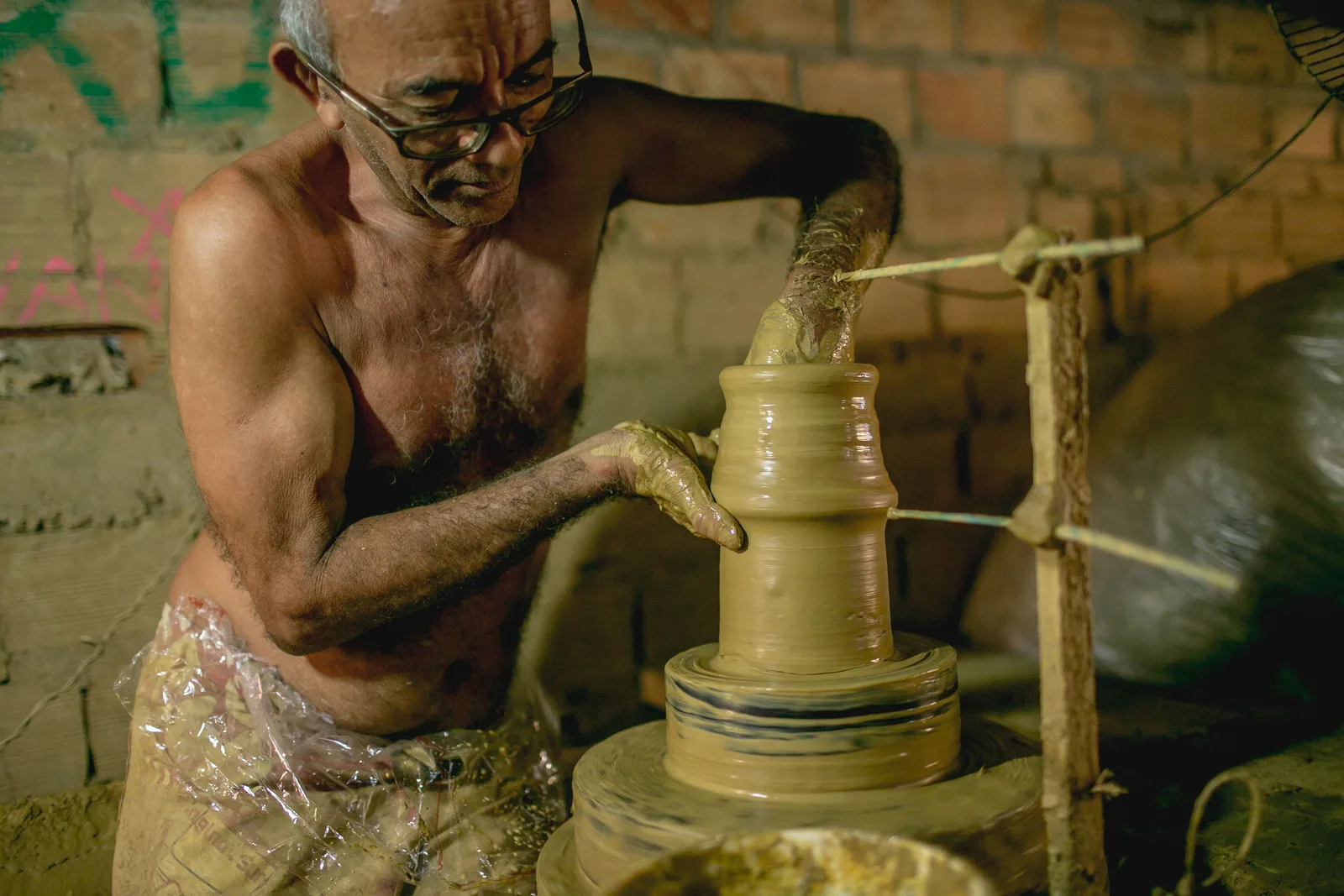 A potter working wet clay into a tall vessel on a wheel in an open brick workshop at Icoaraci