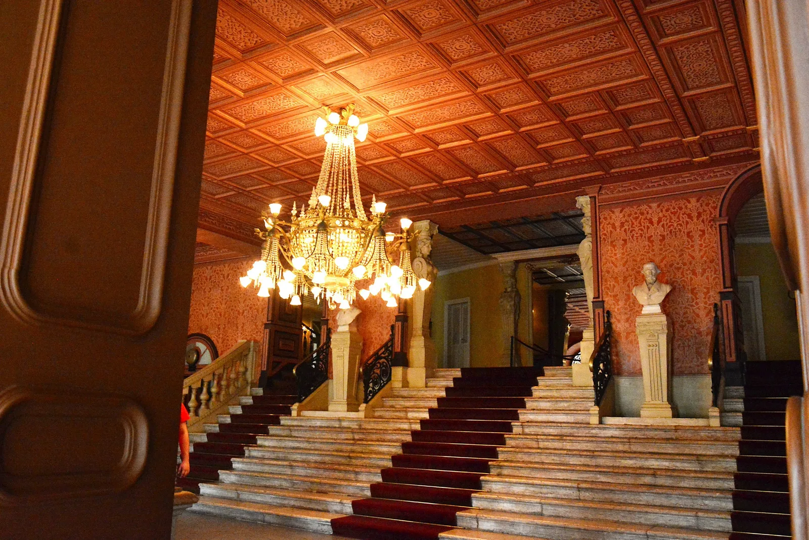 The main staircase inside the Theatro da Paz, red carpet running up between orange walls under a many-armed crystal chandelier