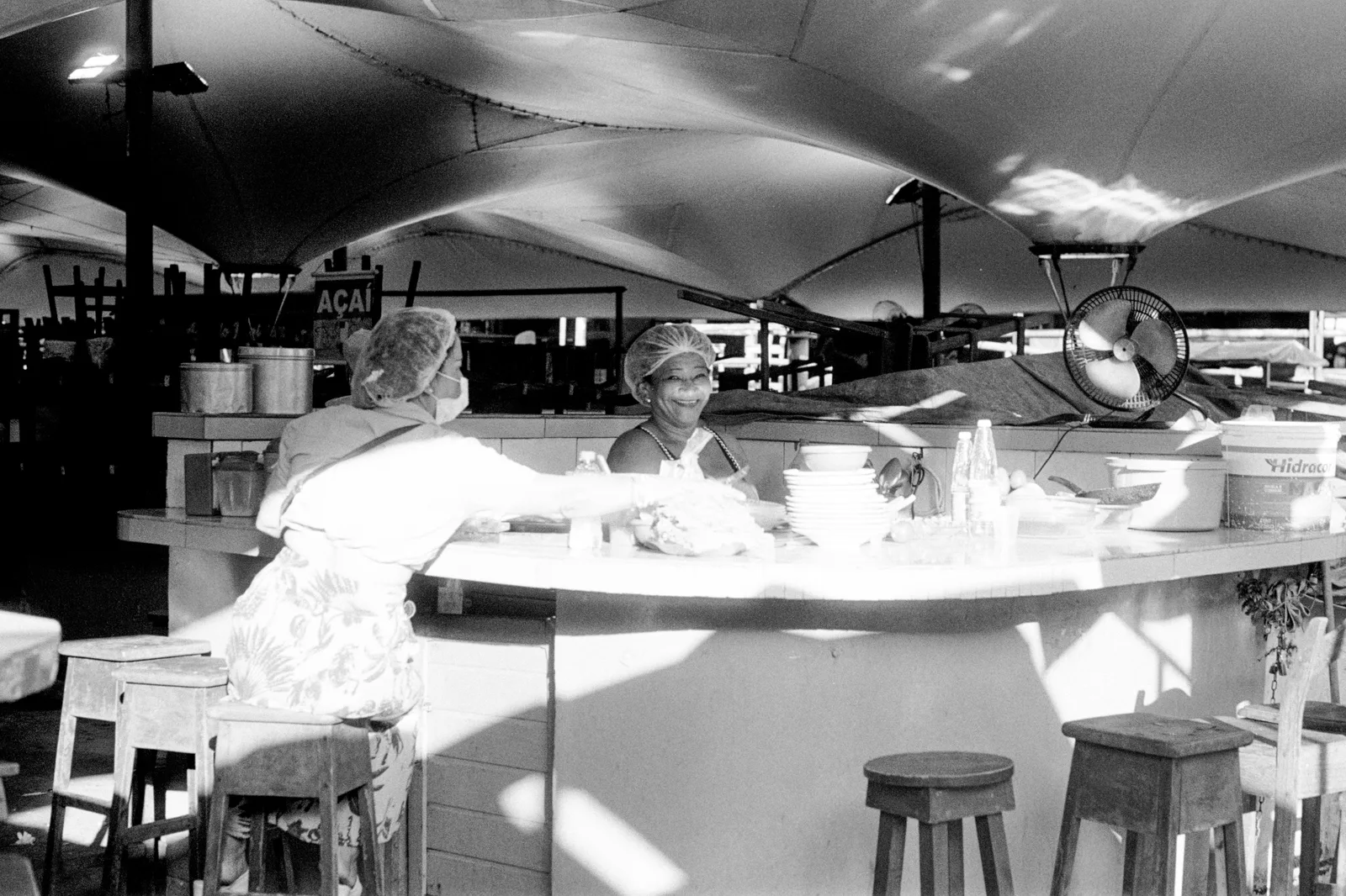 Black and white: two women leaning across a curved food counter under the market roof, pans and dishes stacked in front of them