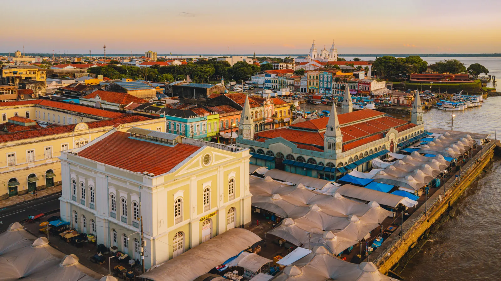 The historic center of Belém from the air in late light: the Ver-o-Peso market square packed edge to edge with white awnings, a long yellow colonial building beside it, tiled roofs running back from the waterfront and the pale bay behind
