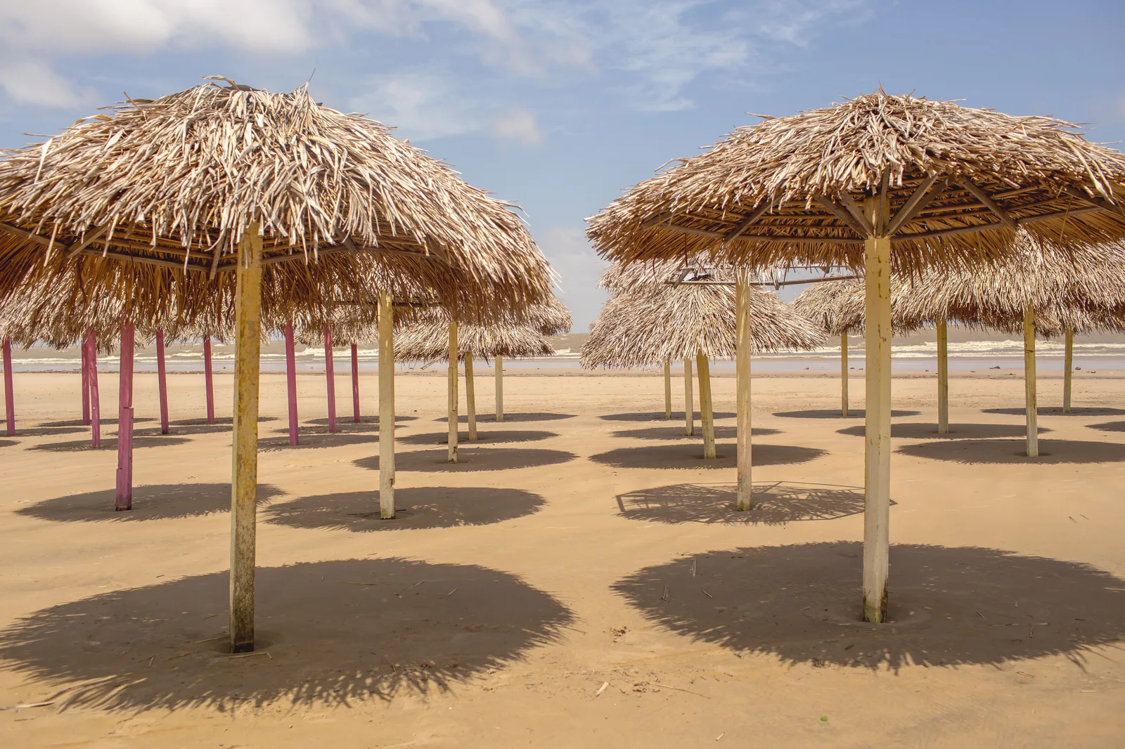Two thatched beach shelters standing on their poles on the wide, empty pale sand at Praia do Pesqueiro on Marajó