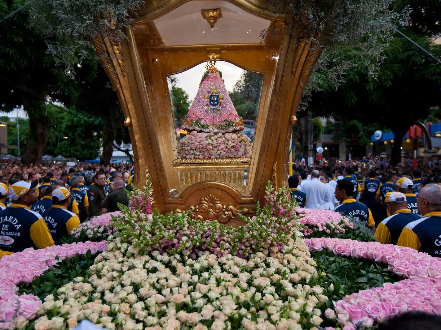 The Círio carriage in the middle of a dense crowd: a gilded canopy over the small robed image of Our Lady, the base banked with pink and white flowers, blue-uniformed guards pressed around it and the street packed solid behind