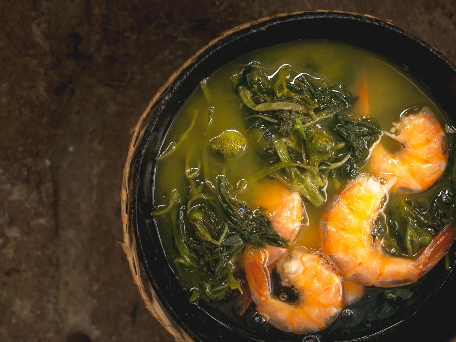 A dark bowl of tacacá seen from above: cloudy yellow broth with three whole pink shrimp and a tangle of dark green jambu leaves floating in it