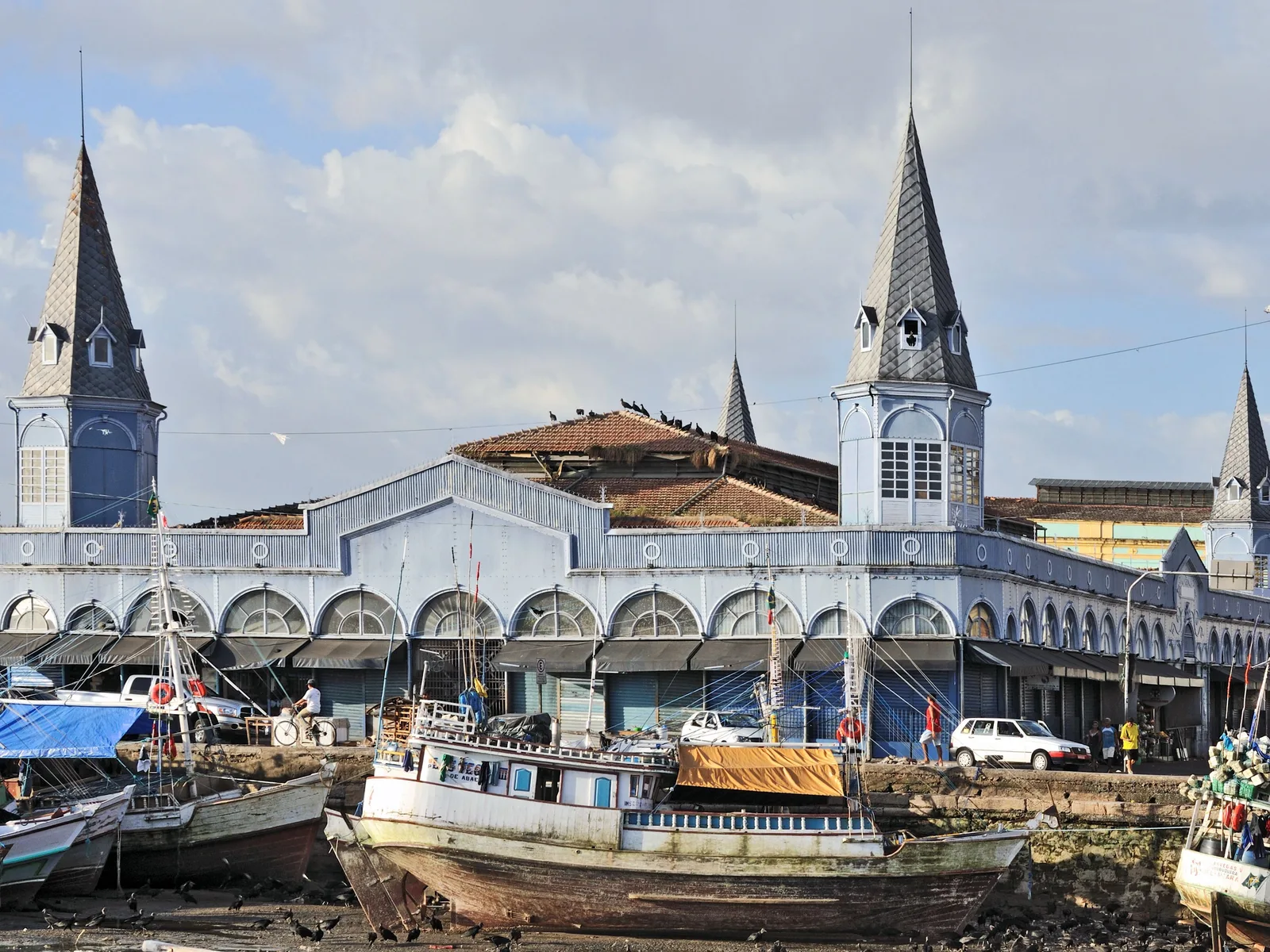 The Ver-o-Peso market from the waterfront: the long white iron pavilion with its four turreted towers under a pale sky, wooden fishing boats hauled up and moored in a line in front of it