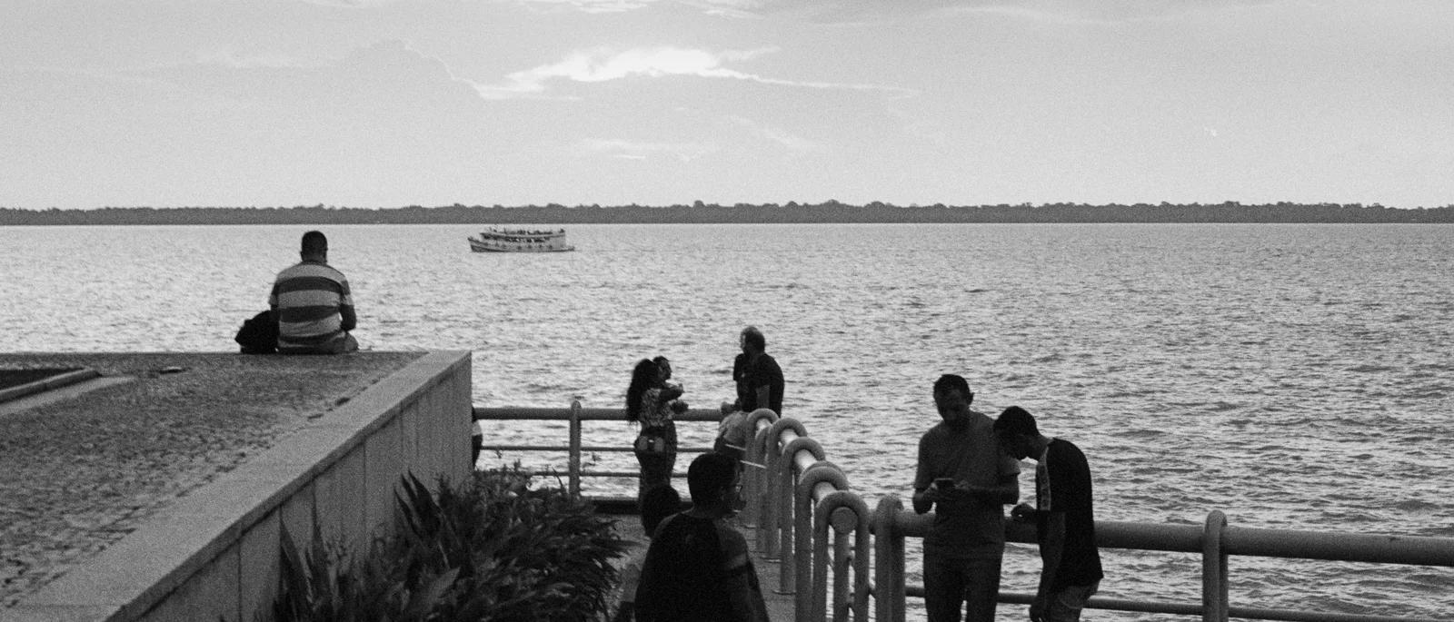 Black and white: people leaning on a waterfront railing in the old city, looking out across the wide bay with a boat on the water in the distance
