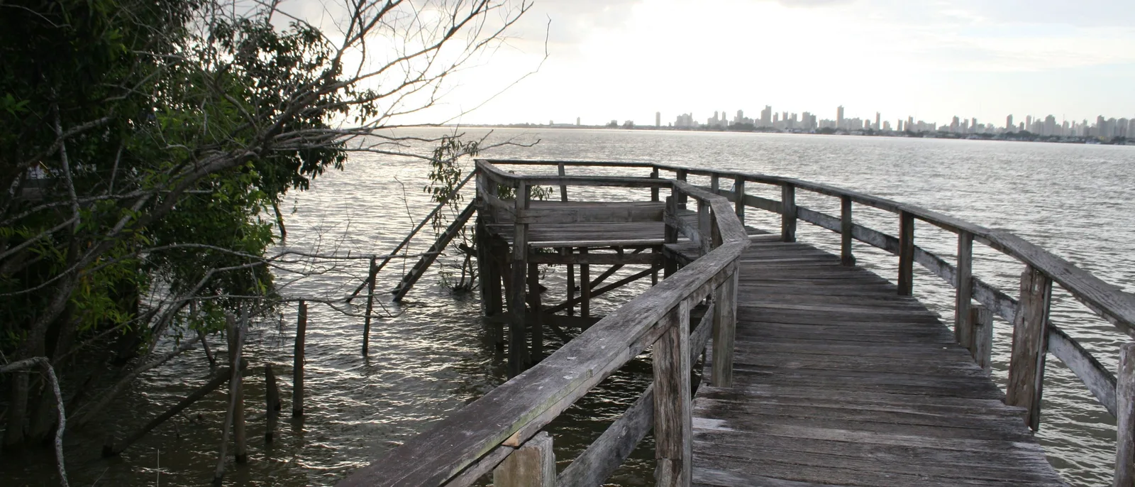 A wooden walkway on posts running out through mangrove toward open brown water, with the distant towers of Belém along the horizon