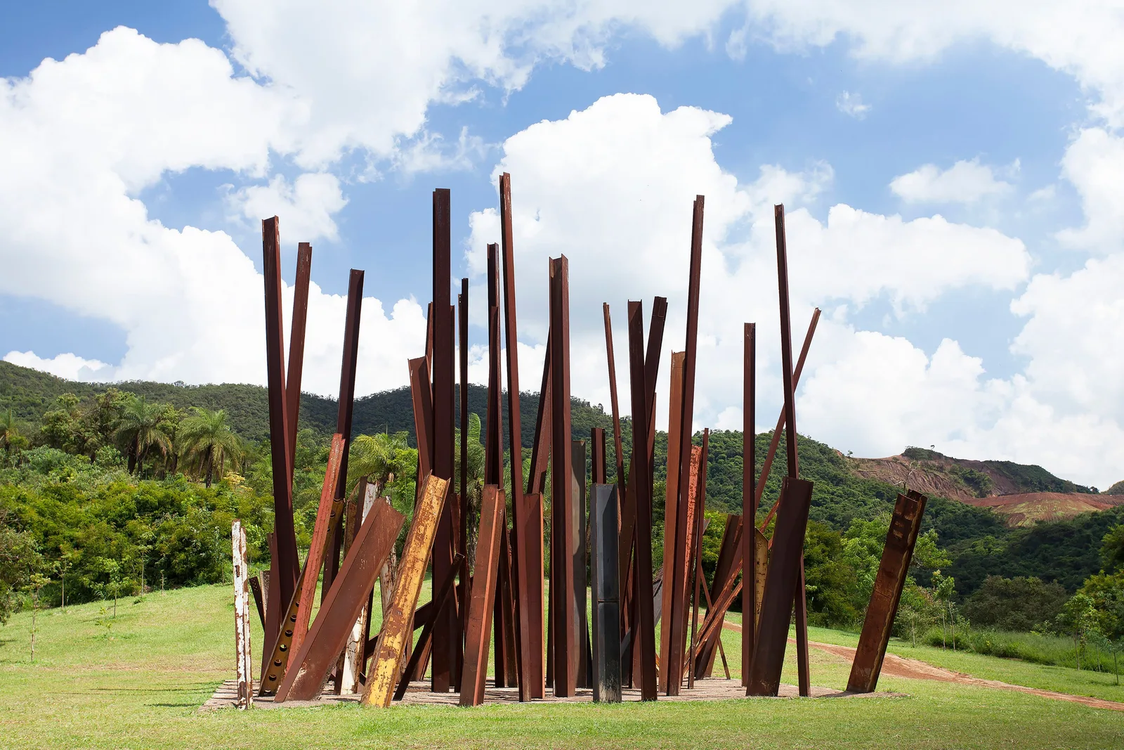 Chris Burden’s Beam Drop at Inhotim: a cluster of rust-red steel beams standing at all angles out of a concrete base on a grassy hilltop, green hills and a reddish mountainside behind under white clouds