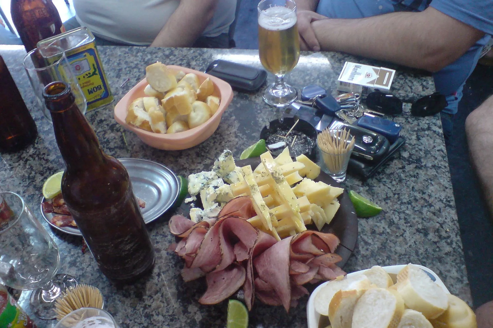 A bar table seen from above with beer glasses and bottles, a board of cheese and sliced ham, bread and lime wedges, friends’ hands at the edge of the frame