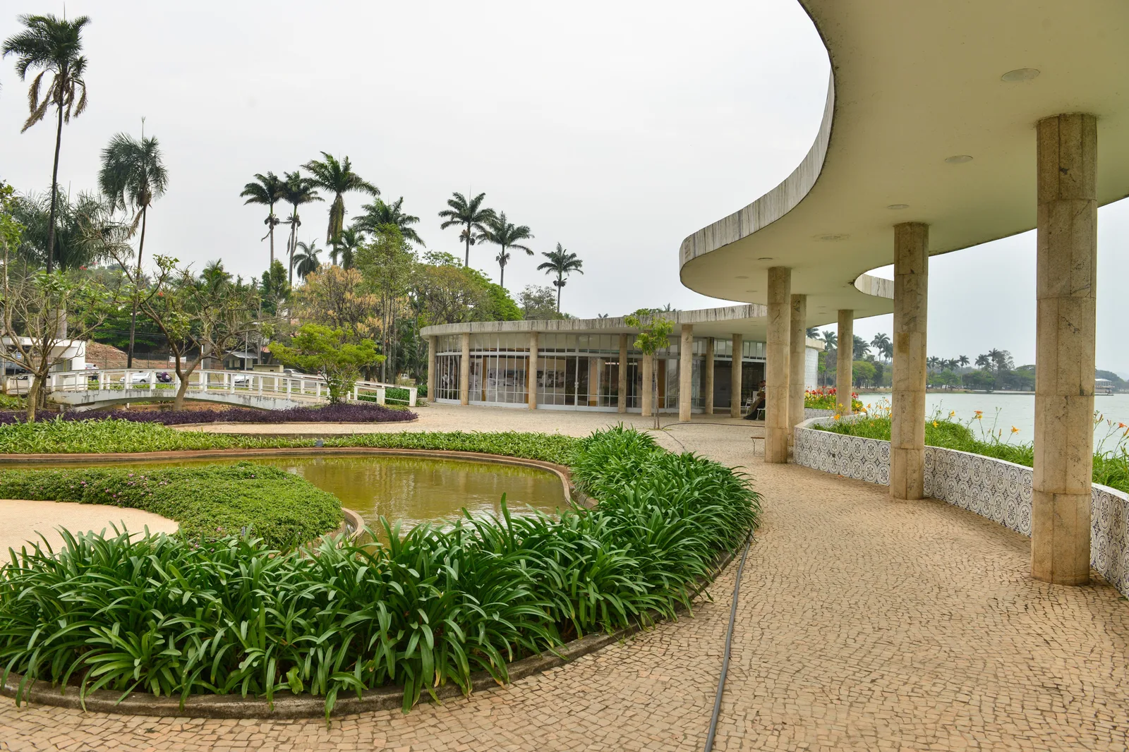 The Casa do Baile at Pampulha: a white concrete canopy on slender columns curving away along a path beside lawns and planted beds, the round pavilion and palms beyond