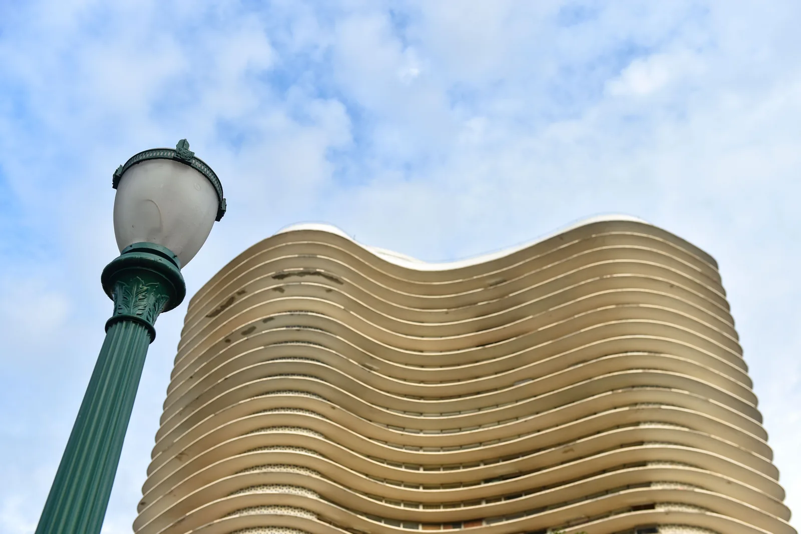 The wavy facade of the Edifício Niemeyer seen from below, its pale horizontal bands rippling across the building, a green iron street lamp in the foreground against a blue sky