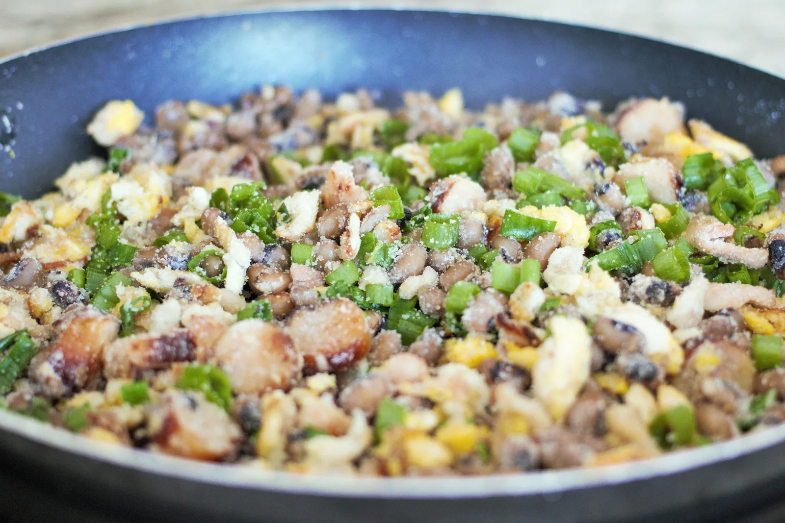 A dark pan full of feijão tropeiro seen from above: beans mixed with toasted manioc flour, scrambled egg and chopped green onion