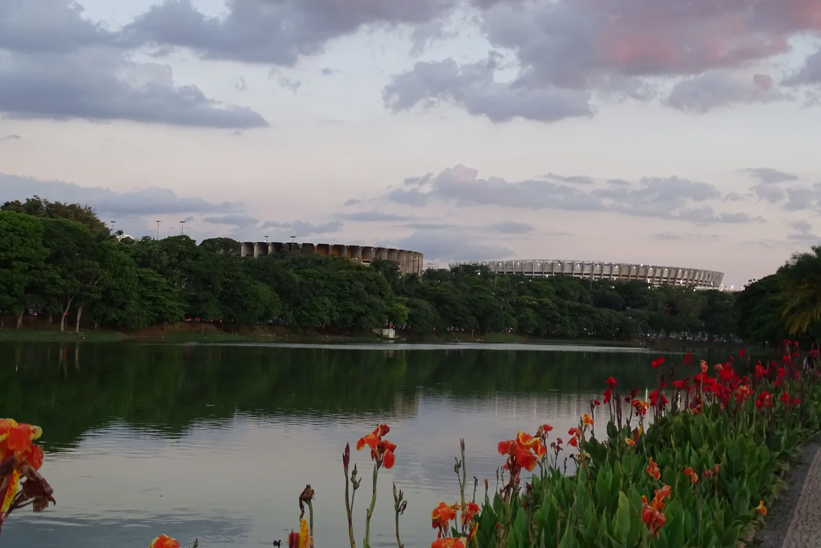 The Pampulha lake at dusk from the shore by the church, orange and red canna flowers along the edge, trees on the far bank and the concrete rings of the Mineirinho and Mineirão stadiums above them under pink-tinged clouds