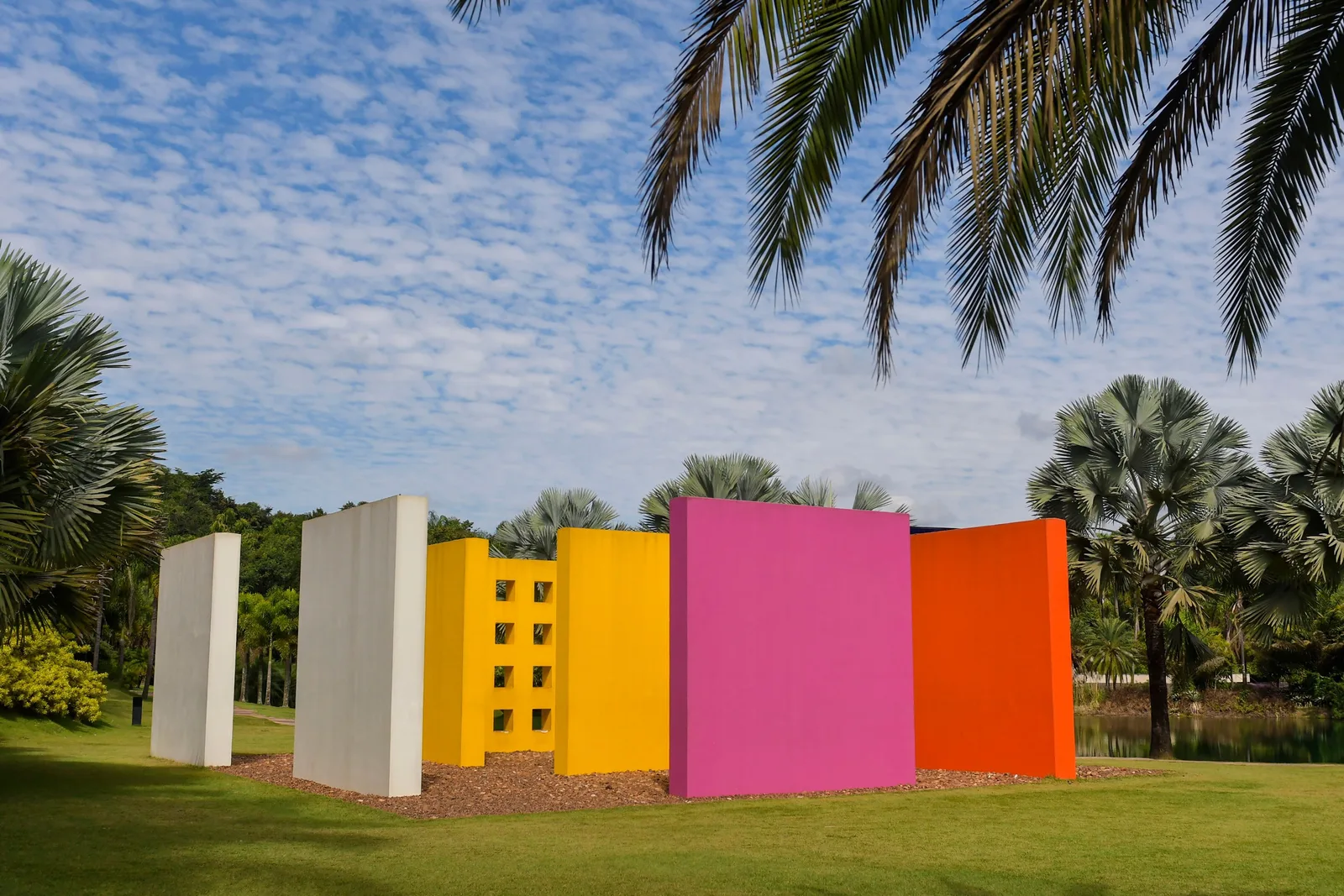 Hélio Oiticica’s Magic Square #5 at Inhotim: freestanding walls painted white, yellow, magenta and orange standing on a lawn under a blue sky, palm fronds hanging into the top of the frame