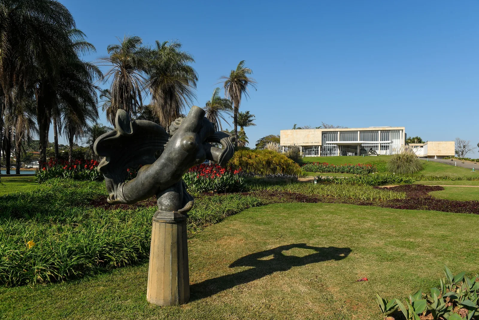 August Zamoyski’s sculpture Nu, dark bronze on a stone plinth, in a garden of lawns and flower beds, palms behind it and the low white modernist building of the Museu de Arte da Pampulha at the far side