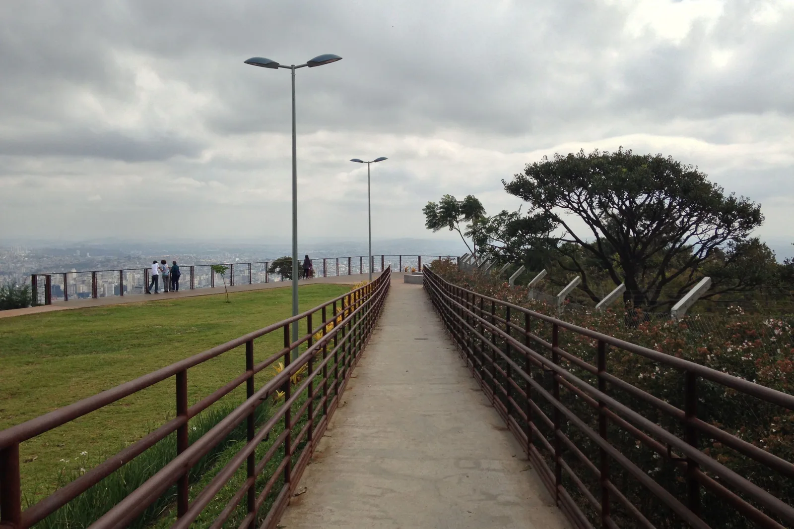 A long railed walkway leading out to a lookout point on a grassy hilltop at the Mirante do Mangabeiras, street lamps along it and a gray sky over the city beyond