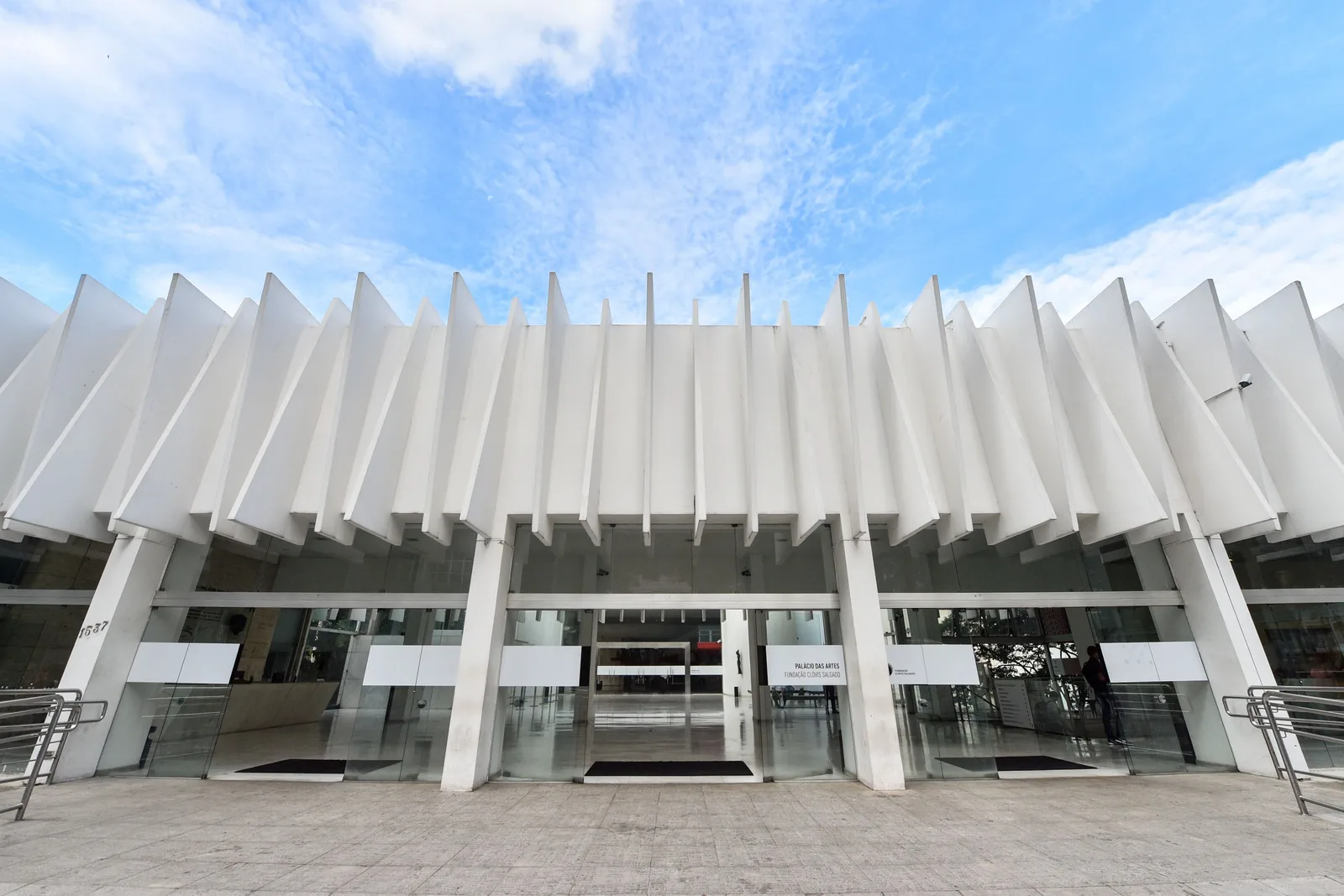 The front of the Palácio das Artes: a long white modernist building with a row of angled concrete fins along its roofline above a glazed entrance, under a blue sky