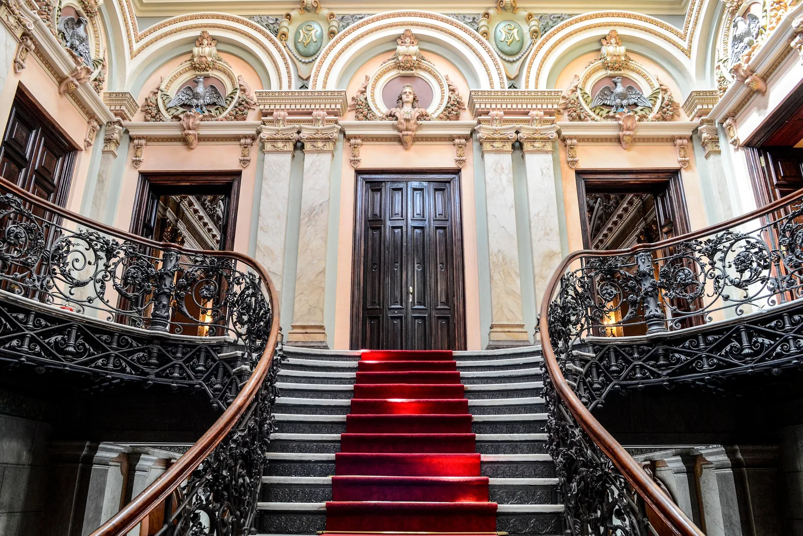 The entrance staircase inside the Palácio da Liberdade: a red carpet running up between two curving wrought-iron balustrades toward a carved wooden door, ornate painted walls above