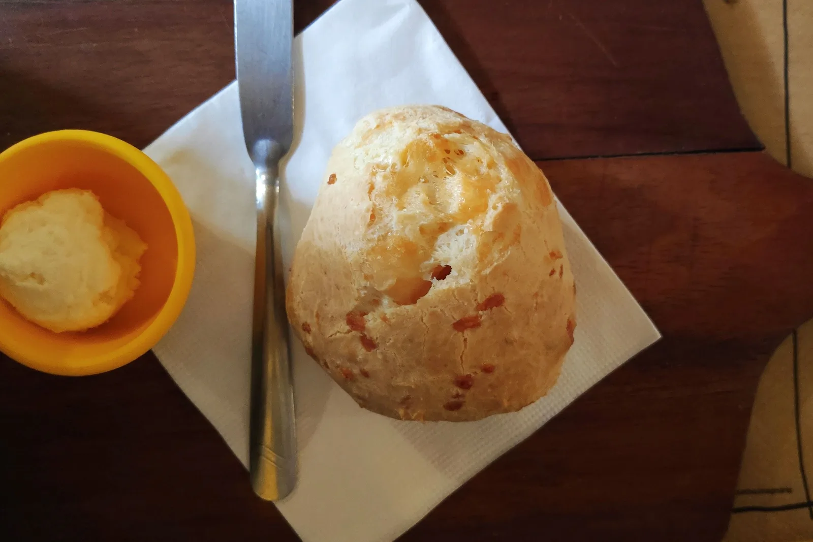 A single golden pão de queijo on a paper napkin beside a small orange dish of soft cheese and a butter knife, on a dark wooden table