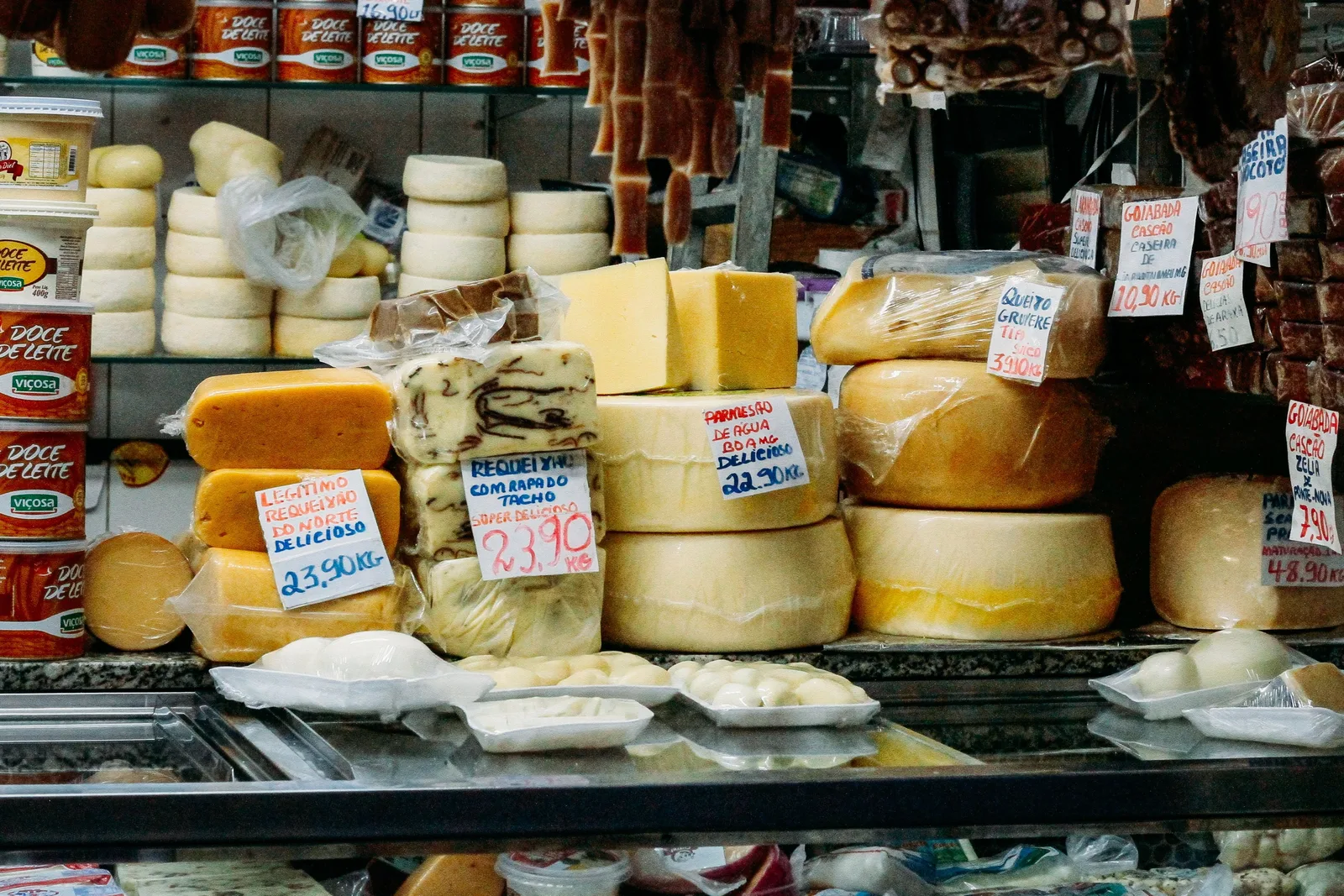 A cheese counter at the Mercado Central piled with whole wheels and wedges of pale and golden cheese, handwritten price signs tucked between them and cured meats hanging above
