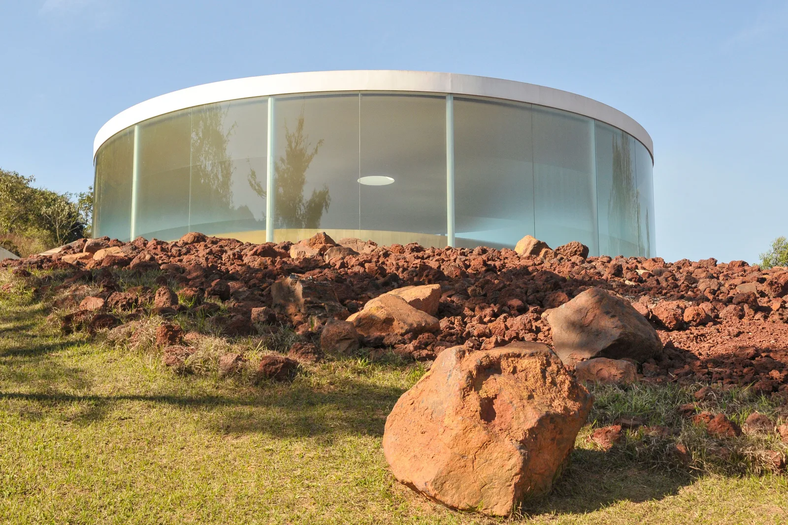 Doug Aitken’s Sonic Pavilion at Inhotim: a low circular pavilion with a white roof and frosted glass walls standing on a hilltop of red earth and scattered boulders under a pale sky