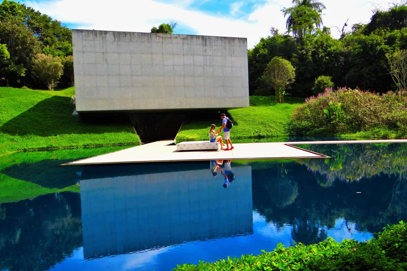 The Adriana Varejão gallery at Inhotim, designed by the architect Rodrigo Cerviño Lopez: a plain gray concrete block raised over a deep blue reflecting pool, a wide white deck at the water’s edge with two visitors on it, green lawns and trees around it